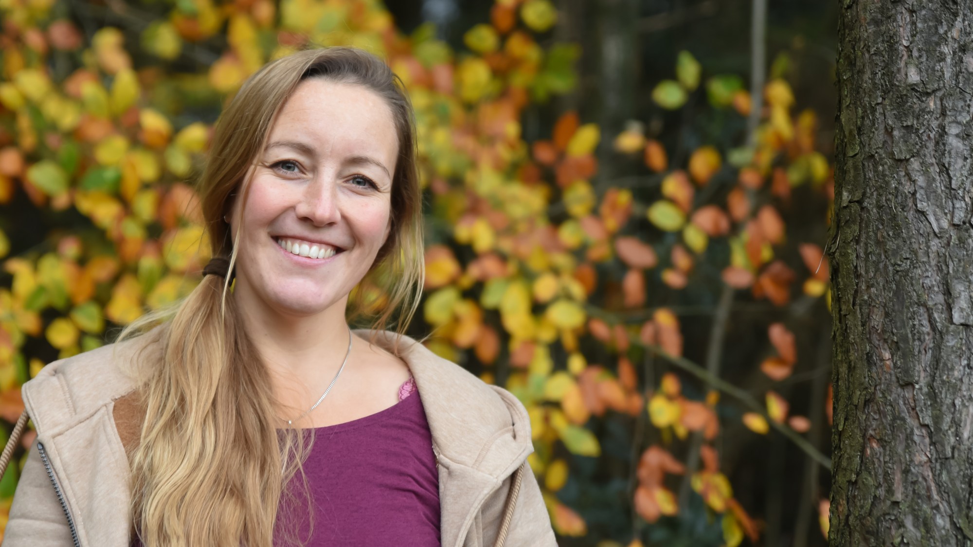 Helke_Lemme_WS4 Helke lacht in die Kamera mit einem bordeauroten Oberteil und einer offenen beigen Pulloverjacke. Im Hintergrund Herbst-Laubwald.