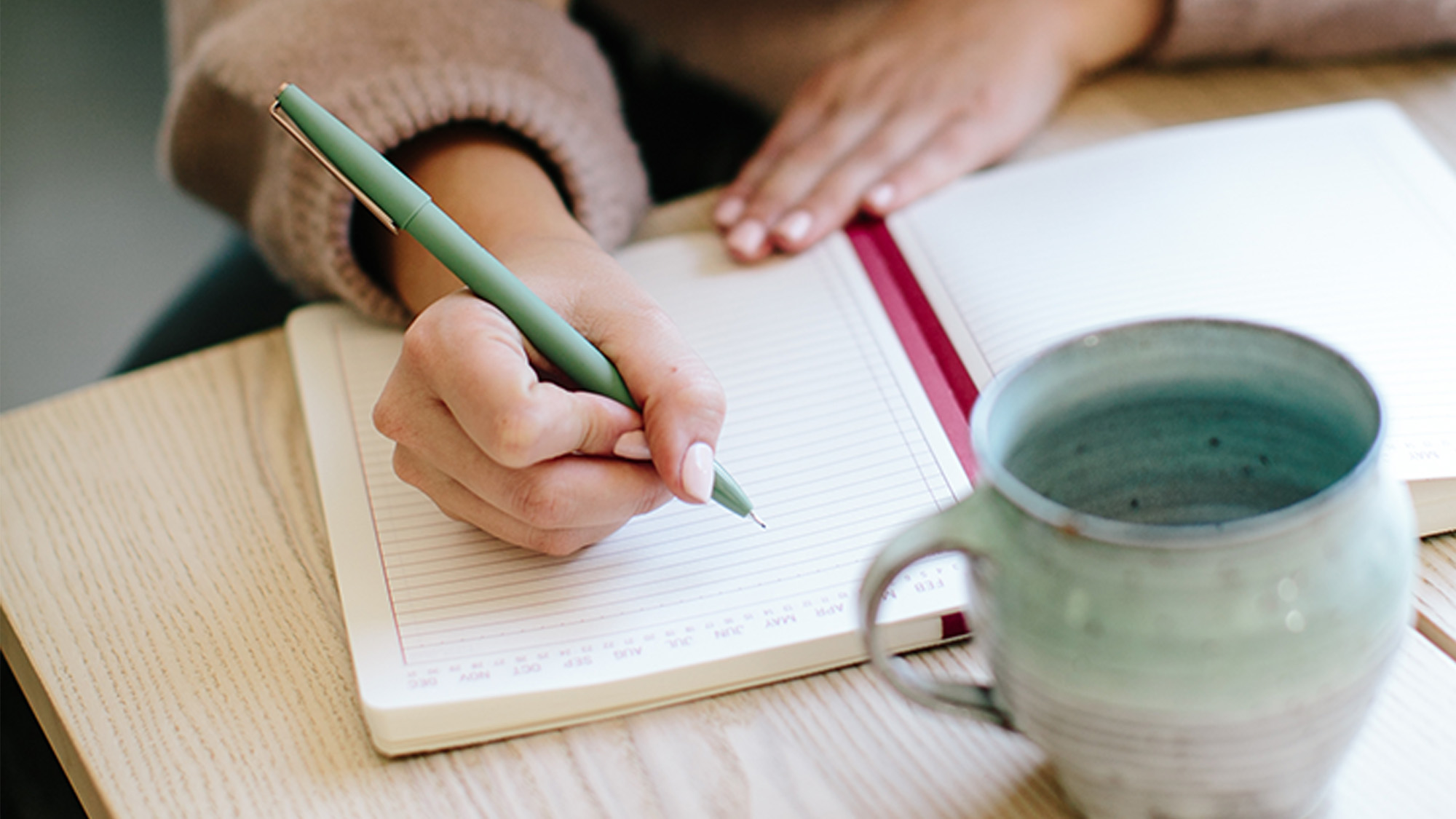Ein aufgeklapptes Notizbuch mit rosa Einband liegt auf einem hellen Holztisch, in das eine Frau mit einem hellgrünen Kuli reinschreibt. Im Vordergrund steht ein grau-grüner Becher.