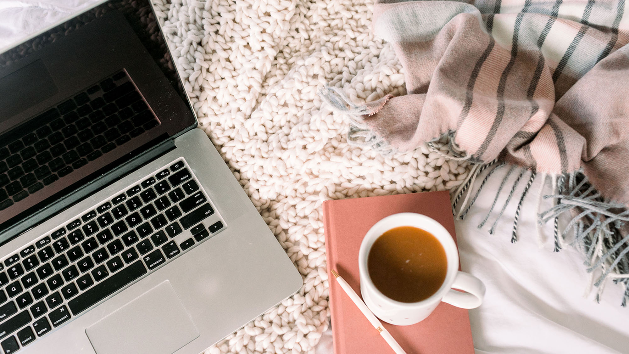 Helke_Lemme_Wohnraum_Reading3 Aufgeklappter Laptop liegt auf einer grobgestrickten beigen Decke. Daneben liegt ein kleines rosa Notizbuch auf dem ein Becher Kaffee und ein weißer Kuli liegt.